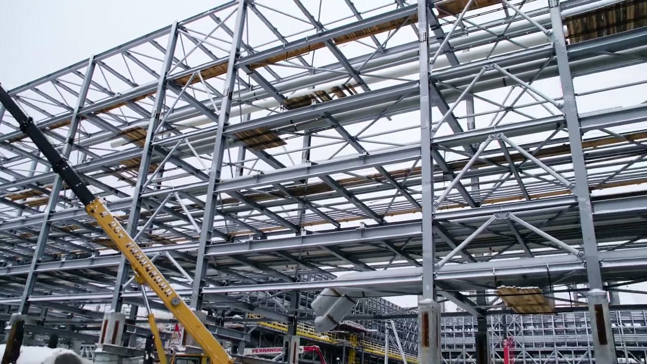 Steel frame erection under flat overcast, with lattice roof trusses, purlins and bracing in place and cladding pending.