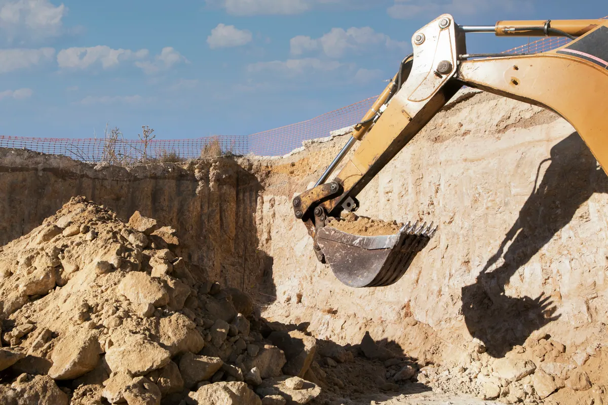 An excavator bucket digging into sandy ground at the edge of a cut.
